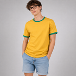 A man wearing a yellow ringer t-shirt with green trim on the collar and sleeve cuffs, paired with light blue denim shorts, standing against a neutral gray background.