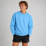 A smiling young man wearing a light blue crewneck sweatshirt and black athletic shorts with a small white Nike swoosh, standing against a light gray studio background.