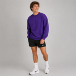 A man modeling a purple crewneck sweatshirt, black athletic shorts with a white Nike logo, white socks, and white Nike running shoes against a solid gray studio background.