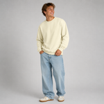 A young man wearing a cream-colored crewneck sweatshirt, light-wash blue jeans, and white sneakers, standing against a neutral gray studio background.