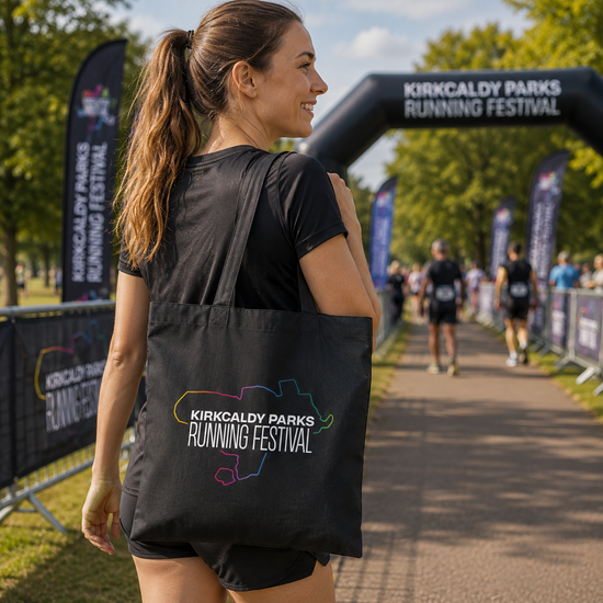A woman wearing a black t-shirt and shorts carries a black tote bag printed with the Kirkcaldy Parks Running Festival logo, walking along a path under a festival arch.