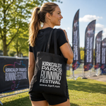 A woman wearing a black t-shirt and shorts carries a black tote bag printed with white text reading 'KIRKCALDY PARKS RUNNING FESTIVAL' and a website URL, photographed outdoors at an event with banners and trees in the background.