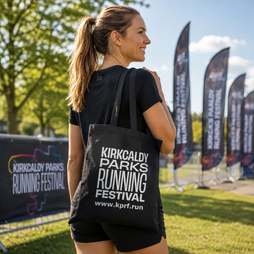 A woman wearing a black t-shirt and shorts carries a black tote bag printed with white text reading 'KIRKCALDY PARKS RUNNING FESTIVAL' and a website URL, photographed outdoors at an event with banners and trees in the background.