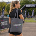 A woman wearing a black t-shirt and shorts is shown from behind, carrying a dark grey tote bag with the 'Kirkcaldy Parks Running Festival' logo printed on it. The background shows an outdoor event setting with banners and trees.