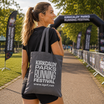 A woman in athletic wear stands on a park path carrying a large grey tote bag printed with the Kirkcaldy Parks Running Festival logo. The background shows event banners, trees, and a running archway.