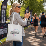 A woman at an outdoor running race carries a white tote bag printed with the Kirkcaldy Parks Running Festival logo. The background shows other runners and event banners on a tree-lined path.