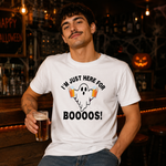 A man wearing a white graphic t-shirt with a Halloween ghost design and the text 'I'M JUST HERE FOR BOOOOS!' while holding a pint of beer in a dimly lit bar setting.