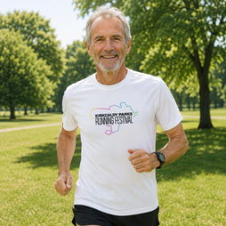 A smiling middle-aged man jogging outdoors in a park wearing a white graphic T-shirt that reads 'Kirkcaldy Downs Running Festival' with a multi-coloured outline map, against a blurred green background.