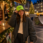 A woman wearing a bright lime green baseball cap with the 'brat Copenhagen' logo, a dark gray bomber jacket, and a cream ribbed sweater, standing in a festive outdoor market at dusk with Christmas trees and lights in the background.