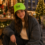 A woman sitting on a wooden bench at an outdoor night market, wearing a bright lime green Brat Company cap, a dark gray bomber jacket, and a cream ribbed sweater, holding a skateboard.
